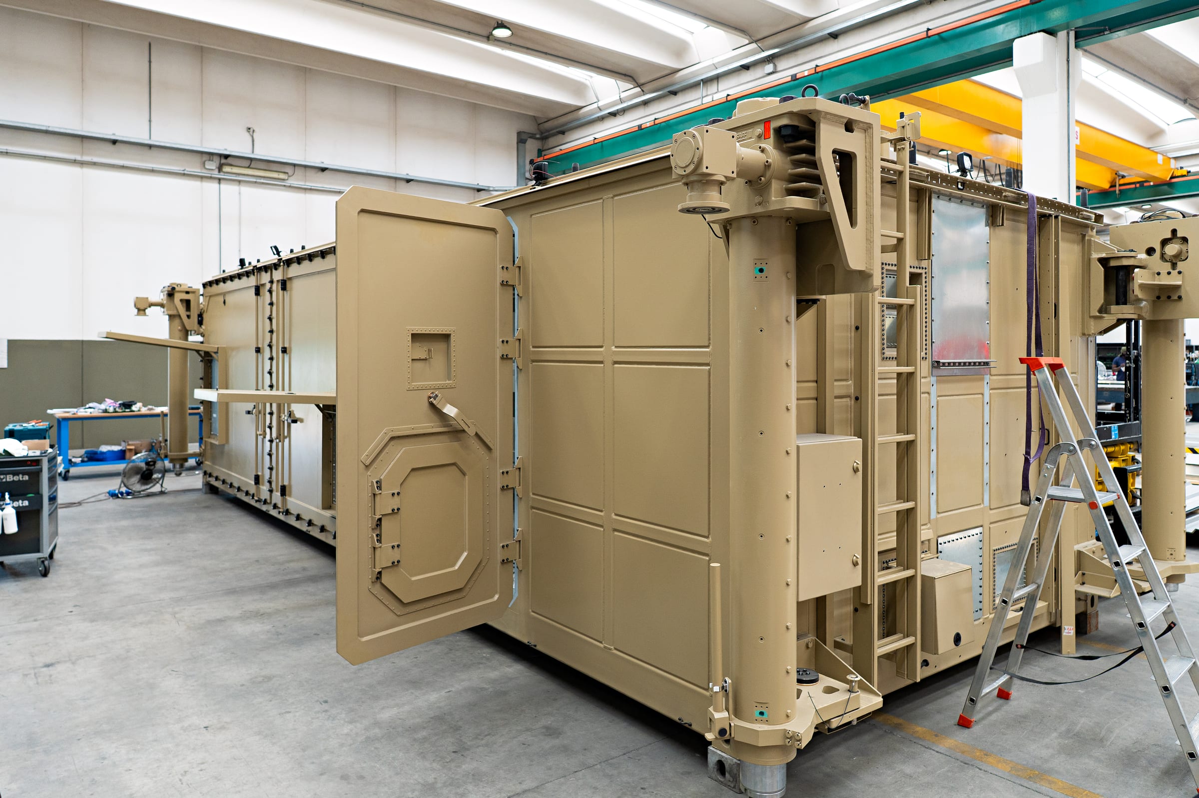 Sand-coloured military shelter with hydraulic legs and open armoured door in the Alex Sistemi hall