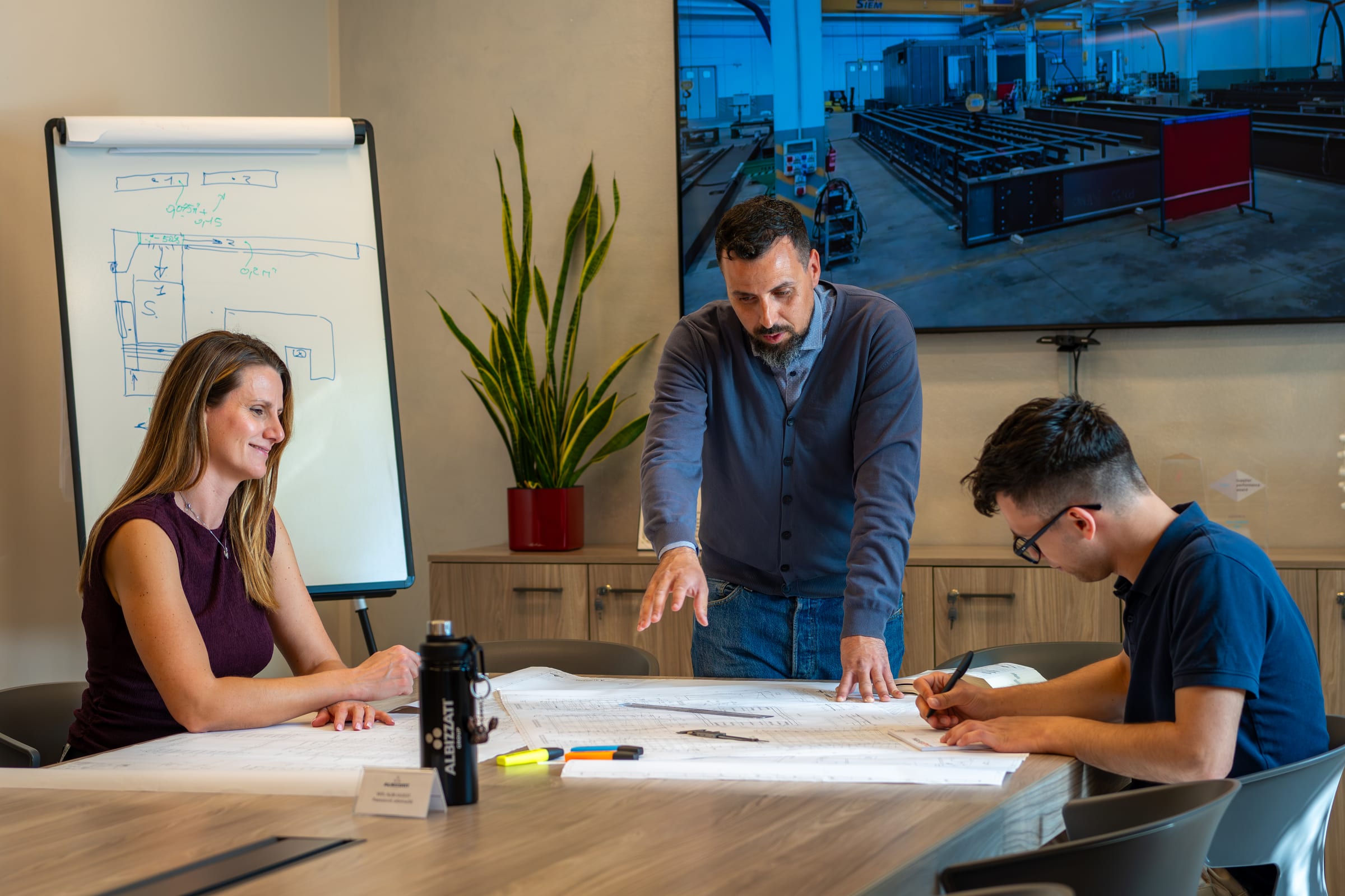Project team reviewing technical drawings around the meeting table, workshop image on the screen